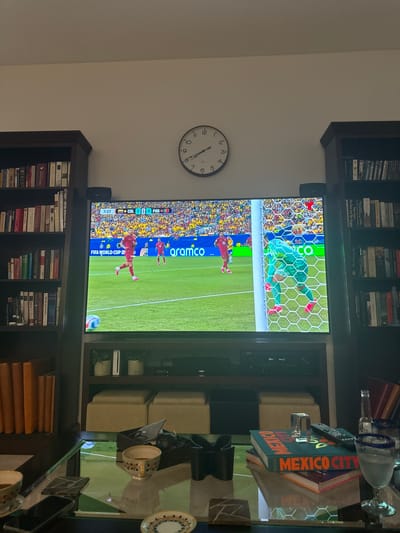 The image depicts a living room scene with a large television displaying a soccer match, likely from the FIFA World Cup, given the "FIFA WORLD CUP" text on the screen. The score shows "COL 0 - 0 POR" with the time at "7:27". Two players in red jerseys are on the field, one near the goal and another further away. A goalkeeper in a green uniform is positioned near the goal. The television is flanked by two tall, dark-wood bookshelves filled with books. Above the television, a round analog clock shows the time as approximately 2:48. In the foreground, a glass coffee table holds several objects, including two cups with coffee, a remote control, a small glass prism, a bottle of Corona beer, and two glasses, one filled with a clear liquid. There are also two books stacked on the table, one with the title "MEXICO CITY" prominently displayed in orange lettering. Behind the coffee table, a media console is visible beneath the television, and two upholstered ottomans are placed in front of it. The overall setting appears to be a comfortable, modern living room. The image was taken in Doral, United States of America.