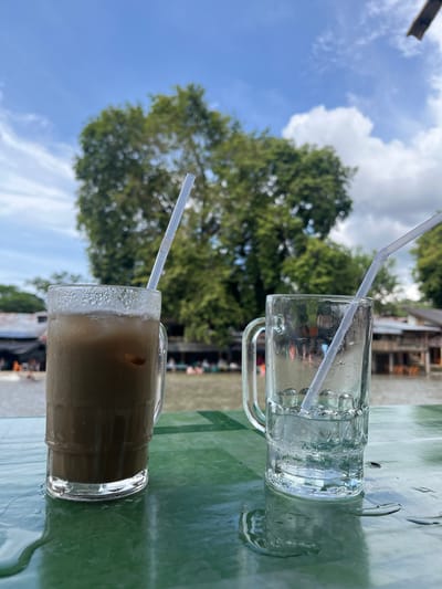 Two iced drinks are placed on a green table in front of a body of water. The drink on the left is a glass filled with a brown liquid and ice. A white straw is in the drink. The glass on the right is filled with ice and water. A white straw is in the glass. In the background, there are trees and buildings. The location is Meurah, Indonesia.