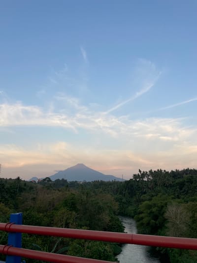 The image depicts a scenic view of a lush green landscape with a mountain in the distance, likely captured from a bridge or elevated vantage point. In the foreground, a red railing with blue supports is visible, suggesting a man-made structure. Beyond the railing, dense vegetation covers rolling hills, with a river winding through the valley. A prominent, cone-shaped mountain dominates the background, silhouetted against a soft, hazy sky with streaks of clouds. The sky transitions from a pale blue at the top to a warm, peachy hue closer to the horizon, indicating either sunrise or sunset. A few scattered buildings and power lines are subtly integrated into the landscape, hinting at human presence in the vicinity. The overall scene evokes a sense of tranquility and natural beauty, characteristic of rural or semi-rural environments. The location is identified as Maumbi, Indonesia.
