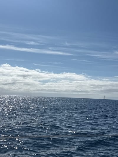 The image displays an expansive ocean seascape offshore from San Miguel de Abona, Spain, under a partially cloudy sky. The upper portion of the frame features a clear light blue sky with horizontal streaks of thin, white cirrus clouds. Below these, a band of denser, white altocumulus clouds stretches across the middle of the horizon. The lower two-thirds of the image are occupied by deep blue ocean water, characterized by numerous small waves and ripples. Intense sunlight reflects off the water's surface, creating a shimmering effect primarily visible in the left and central areas. On the distant horizon, positioned towards the far right, a solitary sailboat with a single white sail is visible. No other vessels or individuals are present in the scene.