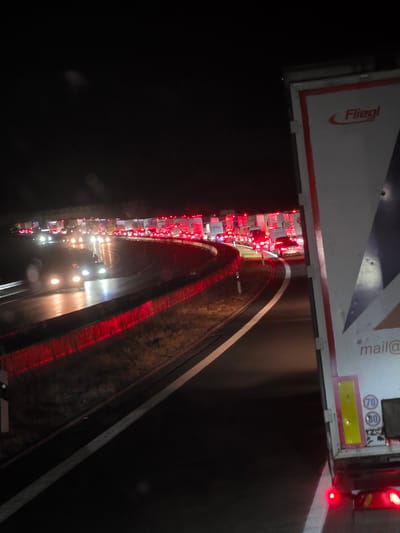The image depicts a nighttime traffic jam on a multi-lane highway, likely near Uettingen, Germany, given the provided information. The road is illuminated by the headlights of numerous vehicles and the red taillights of stationary cars and trucks, creating streaks of light on the wet road surface. The scene is dominated by a large, white semi-trailer truck in the foreground on the right, featuring the "Fliegl" logo. The truck's rear lights are clearly visible and illuminated. In the background, a long line of traffic extends into the darkness, with brake lights and headlights forming a congested pattern. The overall environment suggests a significant disruption to normal traffic flow, possibly due to an accident, road closure, or other event, occurring on a highway that appears to be bordered by grass and possibly a sound barrier or guardrail with red reflective elements. The sky is dark, indicating nighttime conditions.