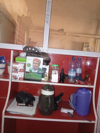 This image depicts a shelf inside a shop in Lankoviri, Nigeria. The shelf is made of red material and is divided into two tiers. On the top shelf, from left to right, are two white cups with blue stripes, a green box, and a framed photograph of a man wearing a red hat and green attire. Next to the photograph are two boxes labeled "T.G. GLOBAL QUICK-ON V80" and a black plastic bag containing an item. On the right side of the top shelf, a yellow box with "SOLAR FAN" is visible.

The lower shelf contains a clear glass jar filled with white powder and a green lid, a small black bottle, two plastic water bottles, one labeled "FARO," a black object that appears to be a speaker, and a small glass bottle with a golden liquid. Further to the right, a dark brown thermos with a white lid is present, along with a black plastic bag containing an object and a pile of papers. A blue plastic jug and a spoon are also on the lower shelf. The overall environment appears to be a retail space, likely a small shop or stall.