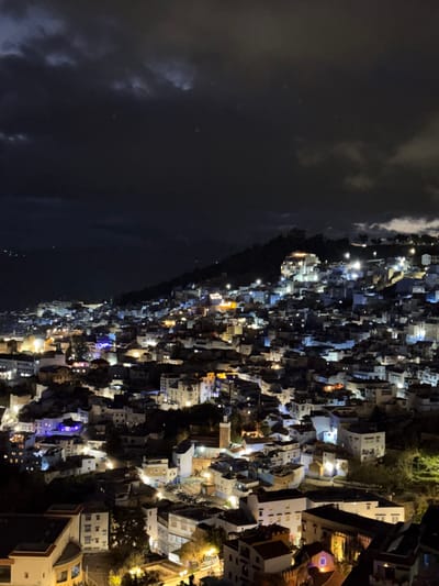 The image displays a panoramic, high-angle nighttime view of the city of Chefchaouen, Morocco, nestled into a steep hillside. The sky above is dark and heavily overcast with visible cloud formations. The city's dense urban fabric extends across the lower and mid-slopes, with buildings illuminated by various light sources. Predominantly, buildings emit warm yellow and orange light from windows and streetlamps, interspersed with distinct pockets of cooler blue and white illumination characteristic of Chefchaouen's architecture. A significant concentration of lights forms a glow around the city, extending upwards along the mountain. A light-brown minaret, belonging to a mosque, is visible in the lower-middle left section of the densely built area. The terrain rises sharply behind the city, with some sparse, dark vegetation discernible on the upper slopes. No individual people, specific actions, or interactions are discernible due to the distance and nighttime conditions. The scene presents a static, illuminated cityscape against a dark, cloudy night sky.
