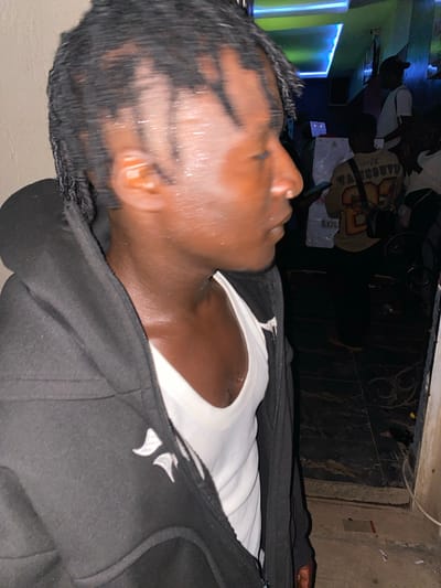 A medium shot captures a person from the chest up, facing right with their head turned away from the camera. They are wearing a black hooded jacket zipped up to the neck, over a white tank top. Their dreadlocks are wet, glistening, and some strands hang over their forehead. The person's skin appears dark and shiny with sweat.
The background is dimly lit and suggests an indoor environment. To the right, several people are visible. One person in the mid-ground wears a yellow shirt with "VANCOUVER 23" on the back. Another person in the background wears a dark cap and a white t-shirt. The environment includes a wall with what appears to be neon blue and green lighting, common in clubs or entertainment venues. A large white object, possibly a cardboard box or display, is also present. The floor is dark and appears tiled in some areas, with a sandy or dusty texture in others. The overall impression is a scene within a lively, possibly a nightclub or bar, setting in Jos, Nigeria.