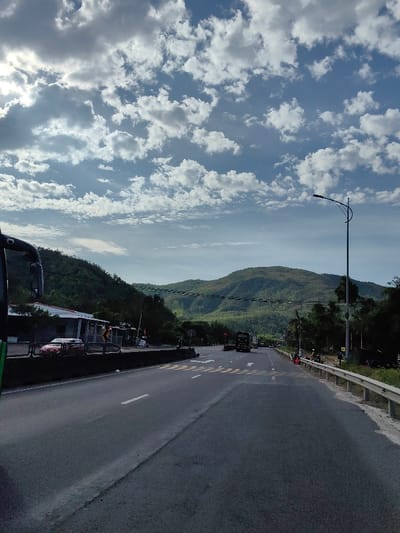 The image displays a multi-lane asphalt road in Hòa Hiệp, Vietnam, under a bright blue sky partially obscured by numerous white cumulus clouds, with sunlight visible in the upper left. The foreground, left, shows a partial view of a dark green vehicle's side mirror. The road features white solid and broken lane markings, along with a chevron pattern on the left side, indicating an intersection or turning area. A red car is parked or stopped off the road to the left, near a light-colored building with a dark roof. Lush green, tree-covered mountains dominate the background. Power lines traverse horizontally across the mid-ground. A tall, dark street lamp with a curved arm stands on the right side of the road. A silver metal guardrail extends along the right shoulder. In the distance, a dark truck or bus is visible in the right lane, traveling away from the viewer, accompanied by other vehicles. Several individuals are gathered on the right shoulder next to motorcycles.