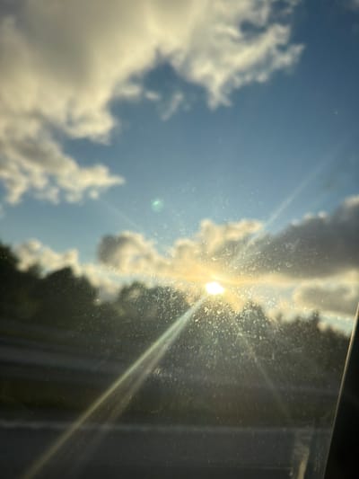 The image depicts a sunlit scene viewed through a dirty window. The sun is positioned in the upper center of the frame, casting bright rays that streak across the image. Clouds, both bright and dark, are visible in the sky, suggesting a partly cloudy day. Below the sky, a silhouette of trees lines the horizon. The foreground is dominated by the window, which is covered in numerous smudges and streaks, obscuring a clear view of the landscape. The overall impression is one of a bright, possibly early morning or late afternoon, sky, seen from inside a vehicle or building. The location is Ödsmål, Sweden.
