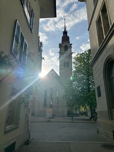 The image presents a sunlit view of a narrow street in a historic urban setting, seen from a low-angle perspective looking upwards.
**Subjects:** The prominent subject is a tall, slender church tower constructed of light-colored stone, featuring a distinctive red-tiled spire topped with a finial and a weather vane. The tower has arched windows and visible architectural details. Flanking the street are older buildings with stone or stucco facades. The building on the left shows multiple windows, some with dark open shutters, and climbing vines on its wall. The building on the right, likely part of the church complex, also exhibits arched windows. The ground is paved with cobblestones. Lush green trees are visible behind and to the side of the church tower, and smaller climbing plants adorn the building on the left. No people are visible in the scene.
**Activity or Event:** No specific activity or event is taking place; the image captures a static architectural street view.
**Notable Details:** The time of day appears to be mid-day or afternoon, characterized by bright sunlight. A prominent sunburst with accompanying lens flares emanates from the sky, positioned between the buildings and the church. The sky is partly cloudy with scattered white clouds. The perspective looks up towards the sky and the towering structures, emphasizing their height. The architecture is indicative of a historic European town.
**Visible Text:** On a sign located on the right side of the street, near the church, the German word "KUNST" (ART) is clearly visible, followed by what appears to be "AUSSTELLUNG" (EXHIBITION). On the facade of the building next to the church, above an arched window, faint text can be observed, including what looks like a name, possibly "O.T. ZWICKY", and a partial date or inscription.