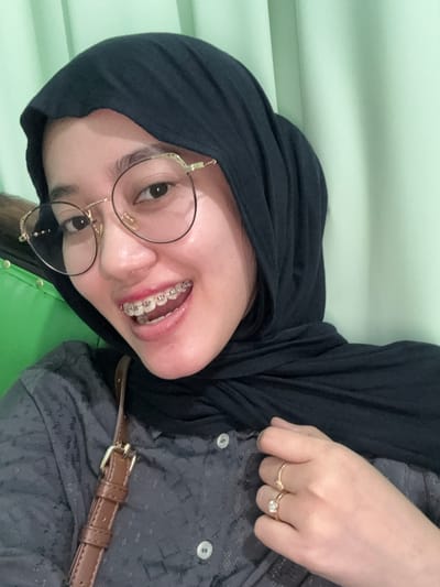 An indoor, close-up, chest-level portrait shot captures a female subject smiling directly at the camera. The individual wears a dark, possibly black, hijab covering her head and neck. She has fair skin and is wearing gold-rimmed, round eyeglasses with decorative accents. Her mouth is open in a wide smile, revealing upper and lower teeth fitted with clear dental braces. She is dressed in a grey, button-up shirt with a subtle patterned texture and light-colored buttons. A brown strap with a gold-toned buckle, possibly from a shoulder bag, is visible over her right shoulder. On the ring finger of her visible left hand, she wears two gold-toned rings: one featuring a prominent clear stone and another simpler band. The background consists of a plain, light mint green wall or curtain, with a darker green object partially visible on the far left. The scene is located in Patrang, Indonesia.