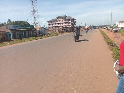 This image captures a street scene in Shirungu-Mugai ward, Kenya, on a clear day. The foreground features a wide, reddish-brown paved road, flanked by dirt and grassy shoulders.
On the left side of the road, a series of single-story commercial structures with corrugated roofs are visible. One prominent building is painted light blue and green, displaying "TSEVAI" and "safaricom m-pesa" signage. Further back, a multi-story building with a pinkish-brown facade and numerous balconies stands out. Behind these structures, a tall, lattice-style telecommunications tower rises, with a dense line of green trees in the distant background. Several individuals are observed near the commercial buildings.
The right side of the road presents a wider, unpaved shoulder with visible tire tracks and muddy depressions. Further down this side, additional buildings, including a white single-story structure, are visible, alongside parked vehicles and more people.
Traffic on the paved road includes multiple motorcycles, specifically "boda-bodas," traveling away from the viewer. At least three motorcycles are clearly discernible in the mid-ground; one rider wears dark attire, another a red shirt. Other vehicles, including a truck, are visible further in the distance.
In the bottom right corner of the frame, the upper torso of an individual wearing a red shirt and blue jeans is partially visible, holding a white motorcycle helmet. The sky is light blue with minimal cloud cover, and the sun casts shadows from the motorcycles towards the left, indicating it is likely mid-morning to early afternoon.