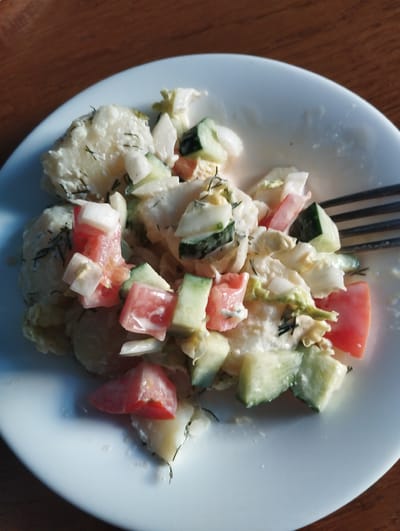 A close-up, overhead shot captures a white plate filled with a potato salad. The salad is a mix of chopped potatoes, diced tomatoes, cucumbers, and white onions, all tossed in a creamy dressing and garnished with fresh dill. A fork rests on the right side of the plate, partially submerged in the salad, suggesting it is being eaten. The plate sits on a dark, wooden surface, likely a table, and the scene is illuminated by natural light, casting soft shadows. There are no people visible in the image. The location is a private residence in Vinkivtsi, Ukraine.