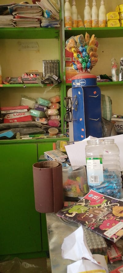 The image displays an interior view of a general merchandise store in Danbare, Nigeria. No individuals are present in the static scene. The store features a light-colored wall background and prominent green wooden shelving units filled with various products.

On the top shelf, stacks of notebooks and miscellaneous stationery items are visible on the left. On the right, six white-capped bottles, possibly beverages with "Sweet..." branding, are arranged alongside yellow packaged goods.

The middle shelf on the left holds stacks of dark-covered books, identifiable as "Oxford" brand, and small items in plastic packaging. Handwritten text is faintly visible on the wall behind these items. The middle shelf on the right features a blue and white NIVEA branded display stand, from which a bouquet of colorful lollipops emerges. A pair of black-framed eyeglasses hangs from a hook on this stand. Below, red and white boxed goods, including one labeled "PERMANENT," and a white bottle with a red cap are visible.

The bottom shelf contains multiple plastic-wrapped packages of colorful, malleable substances, red and white boxes with "LUDE" branding and Chinese characters, and other assorted items.

The foreground reveals a metallic countertop. A large, partially unrolled brown cylindrical object, resembling sandpaper with "P220" text, rests on the left. To its right are two clear plastic jars: a smaller one containing colorful clips, and a larger, ribbed one filled with numerous small blue-wrapped items and displaying a barcode label. A colorful "2A FRIENDS" notebook and crumpled white paper are also on the counter. The overall environment is functional and shows signs of active retail use.