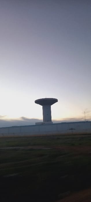 A rotated image displays a concrete structure with a circular top against a sky gradient from blue to pale yellow. The structure appears to be a support or pylon. A landscape featuring green vegetation and a boundary line, potentially a road, is positioned on the right. The image is likely taken during sunrise or sunset, contributing to the soft, diffused lighting and gradient effect.