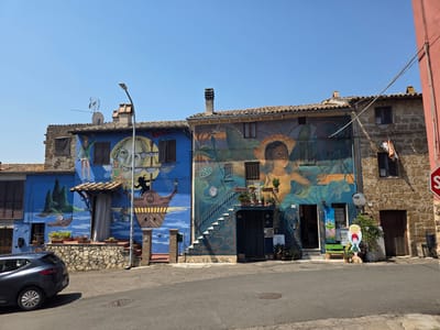 The image shows a street scene in the city of Viterbo, Italy, featuring two buildings adorned with vibrant murals. The building on the left is painted a bright blue and depicts a large face, a sailing ship, and figures in a maritime setting. The building on the right, made of stone, is decorated with a mural of a young woman with flowing hair and subtle mythological or fantastical elements.
In the foreground, a dark grey car is parked on the paved street. A lamppost stands in front of the blue building. The street is lined with a low stone wall and potted plants. Various signs and displays are visible in front of the building on the right, including a cutout of a cartoon character and what appears to be a "For Sale" sign on a window. A red stop sign is visible on the far right edge of the image. The sky is clear and blue, indicating a sunny day. The architecture suggests a historic European town.