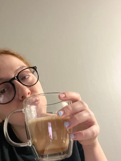 The image displays a close-up, low-angle view of an individual holding and appearing to drink from a clear, double-walled glass mug filled with a light brown, foamy beverage. The person's face is partially visible from the top-left, showing the forehead with reddish-brown hair, dark-framed eyeglasses, and the nose and mouth area. The right hand, with light blue fingernail polish featuring a lighter tip, holds the mug by its handle. The background consists of a plain, light-colored wall. The specific location within Plainsboro Township, United States, cannot be determined from the visual information provided.