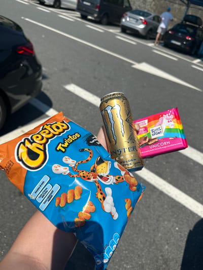 The image shows a person's hand holding a bag of Cheetos Twistos, a can of Monster Energy Golden Apple, and a Ritter Sport Unicorn chocolate bar. The background is a street with parked cars and white lane markings, suggesting a parking area or roadside. The location appears to be in Italy based on the Italian text on the Cheetos package, specifically "Patatine di mais," "Senza conservanti," "Cotte al forno, NON fritte." It cannot be definitively ascertained from the image that the location is Varazze, Italy.