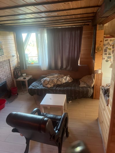 The image shows an interior view of a room with wooden walls and ceiling. The room appears to be a living space or bedroom with a couch covered in a dark patterned duvet and a blanket. There are several pillows on the couch. To the left of the couch, there is a small white table with some items on it, including a box and a device. A fireplace is visible to the far left. In the foreground, a dark leather chair is partially visible. A window with sheer curtains and dark drapes is to the left of the couch. To the right of the couch, there is a white cube-shaped table. Further to the right, a refrigerator is adorned with various magnets and pictures. The floor is made of light-colored wooden planks. The overall impression is of a cozy, somewhat cluttered, rustic interior. The location is identified as Satka, Russia, suggesting this is a room within a dwelling in that city.
