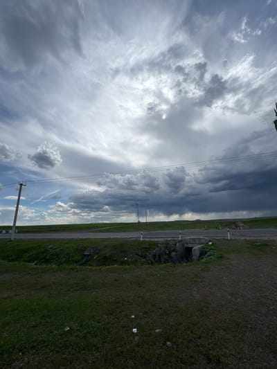 The image shows a landscape in Getk, Armenia, featuring a dramatic sky and a roadside view. The sky occupies a significant portion of the image, displaying a mix of dark, storm-like clouds on the left transitioning to patches of blue sky with scattered white clouds on the right. Power lines stretch across the sky.

A narrow road runs along the lower portion of the image, bordered by grassy terrain. A concrete structure with a small opening is visible near the roadside. Trash is scattered on the grass.