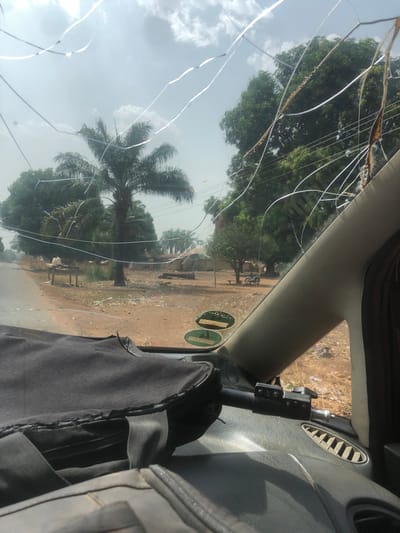 The image shows a view through a cracked windshield of a vehicle, likely taken in Gidan Kado, Nigeria. The outside scene features a dirt road lined with trees, including a palm tree, and scattered buildings in the distance. The sky is overcast. Inside the vehicle, parts of the dashboard, vents, and a bag are visible. There is damage to the windshield due to cracking.