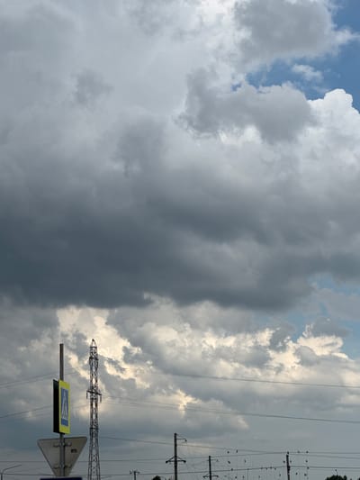 This media file is a vertical, eye-level shot of a dramatic sky over what appears to be an urban or semi-urban environment. The sky is filled with a mix of dark, ominous storm clouds and lighter, brighter clouds that are illuminated by the sun. The contrast between the dark and light clouds creates a sense of impending weather change.
Visible in the foreground on the right side are elements of infrastructure. There are several tall power lines and supporting structures, characterized by a lattice design for the main pylon. Suspended from these lines are numerous small, red flags, possibly decorative or functional. Below the power lines, a pedestrian crossing sign is visible. It is a yellow triangular sign with a blue border, depicting a white pedestrian figure walking. The sign is mounted on a metal pole.
The overall weather appears to be transitioning, with signs of both stormy and fair conditions coexisting. The time of day is difficult to determine precisely due to the overcast nature of some parts of the sky, but the illumination on the brighter clouds suggests it is daytime, likely late afternoon or early evening given the dramatic lighting. There are no people visible in the frame. The setting is likely Rozhdestvenskaya, Russia, as indicated by the location context. The scene evokes a feeling of atmospheric drama and the power of nature.
