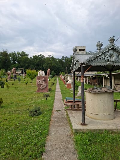 The image depicts an outdoor sculpture park in Malcoci, Moldova. A concrete pathway runs through a grassy area dotted with various statues and busts. On the right side of the pathway, a well structure with a decorative metal roof is prominently featured. The well is made of concrete, and its roof is adorned with intricate metalwork. The park is surrounded by trees and under a cloudy sky. The overall scene conveys a sense of a public space dedicated to art and history.