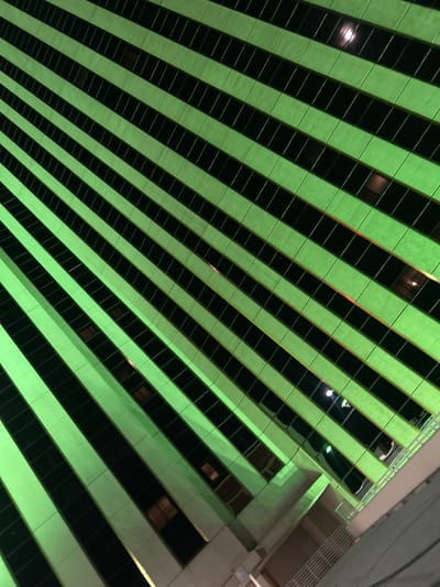 The image presents an upward-angled, nighttime view of the exterior facade of the Peppermill Resort Spa Casino's Tuscany Tower, located in Reno, Nevada, United States of America. The primary subjects are the building's architectural elements, illuminated by artificial light. The facade is characterized by repetitive, diagonal bands composed of alternating sections: bright lime-green illuminated opaque panels and dark, reflective window sections. These bands run consistently from the upper-left to the lower-right of the frame. Some individual window panes exhibit subtle internal or external light reflections. Towards the bottom right of the image, portions of the building's lower structure are visible, including light-colored architectural ledges and sections of white railings. No individuals, discernible actions, or interactions are depicted within the scene. The environment is urban, characterized by modern architecture and artificial night lighting.