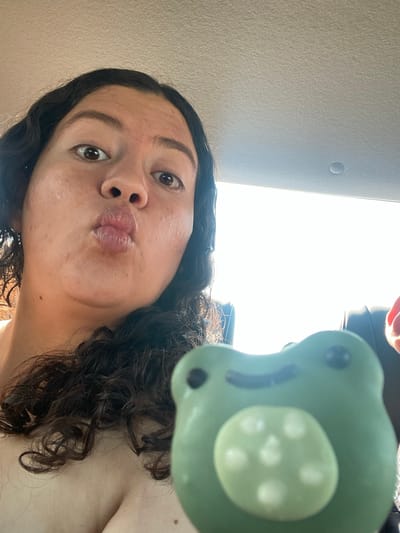 A selfie-style photo appears to be taken inside a car. A woman with curly dark hair and light skin is in the foreground, making a kissing face towards the camera. A green frog-shaped object, possibly a pacifier or toy, is held close to the camera lens and partially obscures the view. The background includes the car's interior, with a light-colored ceiling and a bright light source (possibly the sun) shining through the window. It is impossible to determine the location of Santa Ana Xalmimilulco, Mexico from this image.