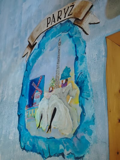 The image shows a painting on a light blue, textured wall, depicting a scene of Paris. At the top of the painting, a banner with the word "PARYŻ" (Paris in Polish) is visible. Below the banner, a stylized frame, rendered to look like rough-hewn rock or ice, encloses a painting of Parisian landmarks. In the center, the Eiffel Tower stands tall, with a stylized representation of the Moulin Rouge windmill to its left. A figure in a wide-brimmed hat and a flowing dress is depicted in the foreground, with legs visible beneath the skirt. The painting is set against a sky-blue background with some white and yellow tones, suggesting a bright day. To the right of the painting, a section of a wooden surface with visible grain and a few small dark spots is present, suggesting the painting is on a wall adjacent to a wooden element. This artistic representation of Paris is located at the Hotelik nad Zalewem, which is situated in the city of Horbów-Kolonia, Poland.