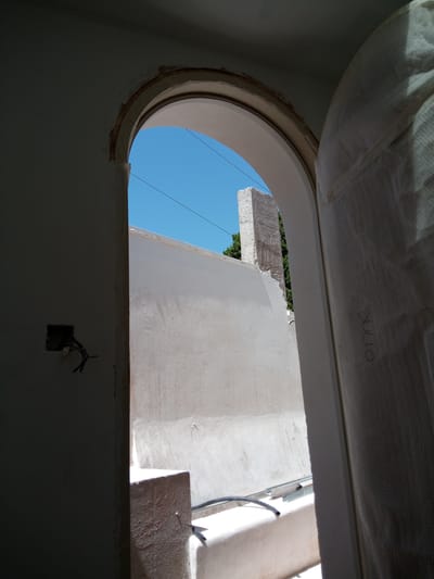 The image depicts an interior view looking outwards through an arched doorway. The doorway is framed by white walls, and the opening reveals a bright blue sky, suggesting it is daytime and clear weather.
Outside, a white-plastered wall forms a sunlit courtyard or patio. A section of a tall, rough-textured, rectangular structure, possibly a pillar or chimney, is visible against the sky. Green foliage peeks out from behind this structure, indicating the presence of trees or bushes.
In the foreground, on the left wall inside the doorway, there is an electrical box with exposed wires, suggesting ongoing construction or renovation. To the right of the doorway, a large, white material, possibly a protective covering or insulation, is draped, obscuring part of the view. Handwritten text, "SULLO", is visible on this white material.
The scene appears to be in a Mediterranean setting, given the bright sunlight, blue sky, and whitewashed architecture common in regions like Nueva Andalucía, Spain. The overall impression is one of an unfinished or partially completed construction site bathed in natural light. There are no people visible in the image. The focus is on the architectural elements and the transition from the interior to the exterior.