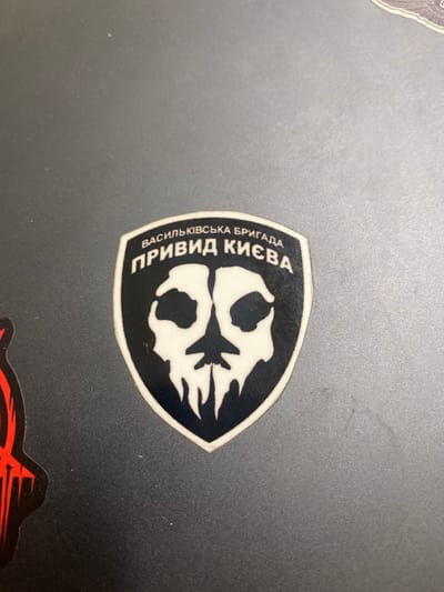 A black and white sticker is affixed to a grey surface. It depicts a skull with a beard. The sticker is shaped like a shield and bears the inscription "Васильківська бригада Привид Києва" in Ukrainian. This sticker is likely located in Richmond, Virginia, based on the provided coordinates.