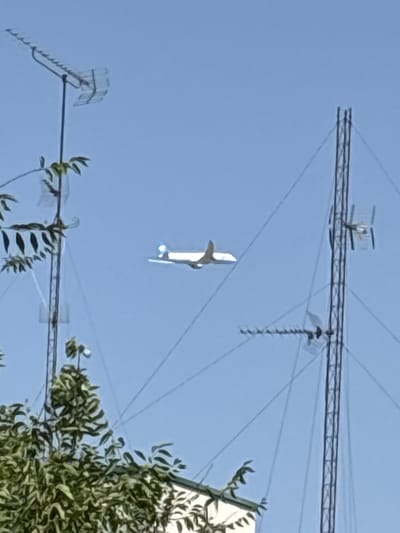 The image shows a white model airplane with blue accents flying in a clear blue sky. The airplane is in flight, with its wings and tail visible. In the background, there are several large antenna structures, likely part of a radio or communication tower. Some of these antennas have a grid-like or comb-like appearance, with horizontal and vertical elements. There are also several thin wires crisscrossing the sky, connecting the antennas or supporting them. To the right of the image, a portion of a building with white siding is visible, partially obscured by lush green foliage. The overall scene suggests an outdoor setting, possibly during the daytime due to the bright sky and clear visibility. The presence of the antennas and the model airplane could indicate a location near an airport, a communication facility, or a place where hobbyists fly remote-controlled aircraft. No people are visible in the frame, and the image does not convey any specific emotion. There is no visible text in the image.