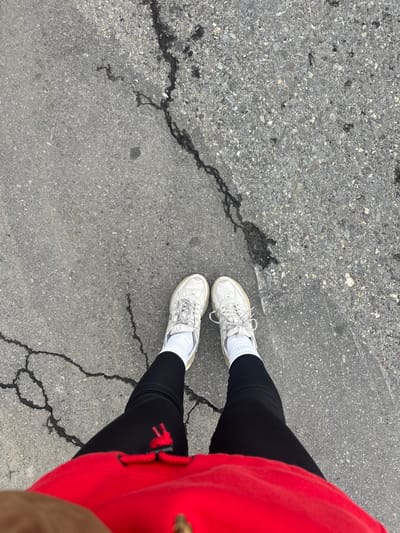 A first-person perspective photograph shows a person standing on cracked asphalt, potentially in Nicolosi, Italy. The individual is wearing white sneakers with white socks and black leggings. A red hoodie is partially visible on the right side of the image. The asphalt surface exhibits a network of cracks and variations in texture. No identifying elements like street signs or landmarks are visible. No interactions between the person and the environment can be interpreted from the image.