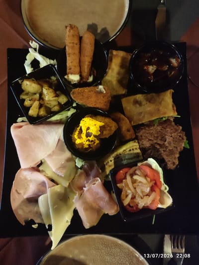 A close-up overhead shot shows a black platter filled with various appetizers, likely part of a tasting menu or antipasto. The platter is placed on a brown tablecloth. In the upper left is a portion of a large, round, beige ceramic plate with a textured rim.
The main platter contains several distinct food items arranged symmetrically. In the top row, there are two golden-brown fried mozzarella sticks, each with melted cheese oozing out, served in a small black bowl. To their right, a black bowl holds a dark, rich sauce, possibly a reduction or barbecue sauce, with small pieces of meat submerged. Beside these, to the left, is a small black square dish filled with roasted potato wedges seasoned with herbs. Below the mozzarella sticks, a rectangular fried item, possibly a spring roll or savory pastry, sits on the platter, partially covering a crispy, breaded food item, likely another appetizer.
The lower portion of the platter features a selection of cured meats and cheeses. Slices of pink ham and white cheese, possibly provolone or mozzarella, are layered and folded. Below these, to the right, is a small black square dish containing a salad of sliced tomatoes and thinly sliced onions, garnished with lettuce. Between the cured meats and the tomato salad, there are grilled slices of zucchini and a dense, dark brown patty, resembling a burger or a meatball, partially covered by a folded pastry.
In the bottom right corner of the image, a fork and knife are visible, along with a timestamp that reads "13/07/2026 22:08". The scene is well-lit, highlighting the textures and colors of the food. The overall impression is of a carefully arranged and appealing presentation of diverse appetizers. There is no specific indication of the location within the city of Scordia, Italy, beyond the possibility that this is a restaurant serving local cuisine.