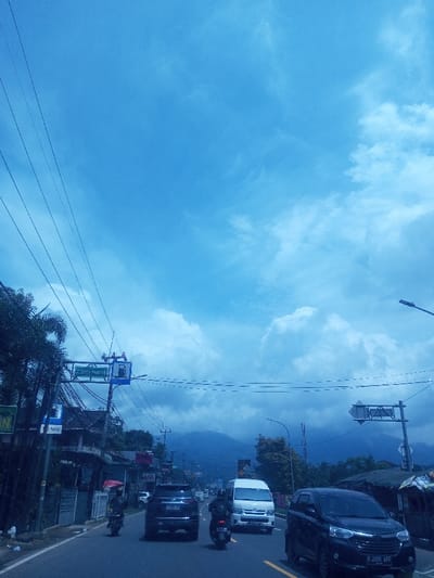 This image depicts a street scene in the city of Chori, Nigeria, under a bright, partly cloudy blue sky, with dark mountainous terrain visible in the distant background. The foreground shows an asphalt road, appearing to have at least two lanes of traffic.
On the left side of the road, a dark-colored SUV is observed, its rear license plate partially visible. Ahead of the SUV, a person on a motorcycle is driving while holding a red umbrella. Further down the left lane, a white passenger van is visible, followed by another motorcycle. In the right lane, a dark minivan is present, also with its rear license plate partially visible.
The left side of the road is bordered by a series of low-rise buildings with various roof types, interspersed with green trees and dense foliage. Multiple utility poles line this side of the street, supporting numerous power lines that crisscross the upper left portion of the frame. A prominent blue street sign, attached to one of the utility poles, features white text and a gas pump icon. On the right side of the road, less defined structures, possibly vendor stalls or small shops, are partially obscured by trees and the passing minivan. The scene captures an active urban or semi-urban environment during daylight hours.