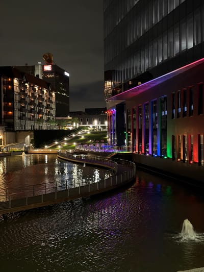 At night in Maryland Heights, Missouri, a modern building with large glass windows is illuminated with colored lights, creating reflections in the water below. A walkway curves along the water's edge, with railings and lights. Across the water, a building with a traditional architectural style is lit, and beyond that, a tall skyscraper is visible with a large, orange, balloon-like structure on top. A fountain sprays water in the foreground, and the water surface is rippled and reflects the ambient lighting. The sky is dark and cloudy.
