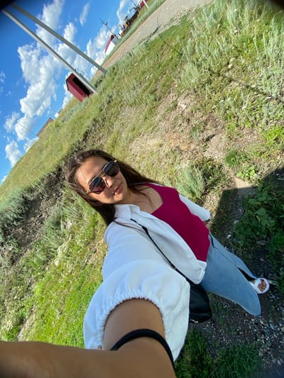 A woman, wearing sunglasses, a fuchsia tank top, a white button-up shirt, and light blue jeans, is taking a selfie outdoors on a sunny day. She is standing on a grassy, hilly terrain with scattered rocks and dry vegetation. In the background, there is a gravel road, a red building, utility poles, and distant industrial equipment. The sky is blue with scattered white clouds. The scene appears to be in a rural or semi-industrial area.