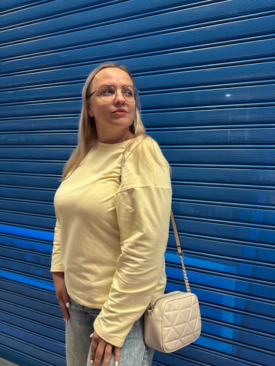 A woman with blonde hair and glasses stands in front of a blue, corrugated metal wall. She is wearing a pale yellow long-sleeved shirt and grey jeans, with a light-colored quilted crossbody bag. The woman is facing to her right, looking away from the camera. The lighting appears to be artificial, casting a soft glow on the scene. The location is not identifiable as Ermakovo, Russia.