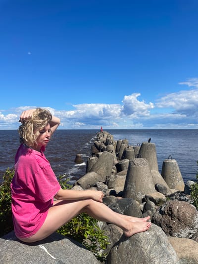 A young woman with blonde, curly hair, wearing a bright pink t-shirt dress, sits on a large rock facing away from the camera, looking out at the sea. Her right hand is raised, touching her hair, and her left arm rests on her lap. She is barefoot, with her left leg extended and her right leg bent. The sea is a deep blue-grey, and a breakwater made of large concrete tetrapods extends into the water, with a small red buoy visible in the distance. The sky is a bright blue with scattered white clouds. The woman is situated in the foreground, with the breakwater and sea occupying the midground and background. The setting appears to be a coastal area, possibly near Belozersk, Russia, given the presence of a breakwater and the general appearance of the landscape.
