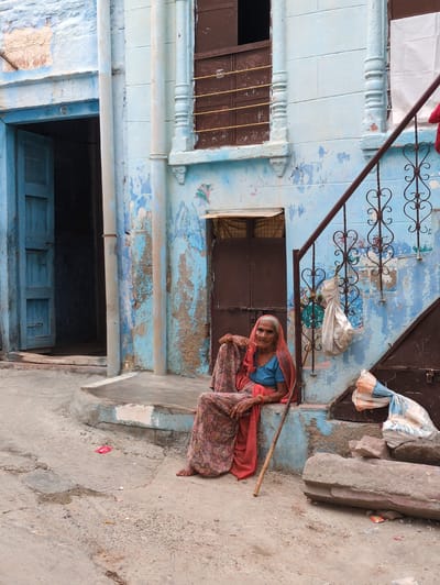 An elderly South Asian woman sits on the steps of a weathered blue building in Jodhpur, India. She is dressed in a patterned skirt, a blue blouse, and a pink headscarf. Her right hand rests on her lap, and her left hand is propped up on a wooden stick. The building's facade is peeling and shows signs of age, with patches of exposed concrete and dirt. A dark wooden door is visible to her left, and a metal staircase with intricate scrollwork ascends to her right. Two white bags are hanging from the staircase railing. The ground in front of the building is a rough, uneven surface of concrete and dirt, with some scattered debris, including a small red object. The overall scene depicts a common sight in the older parts of the city, characterized by its vibrant colors and rustic architecture.