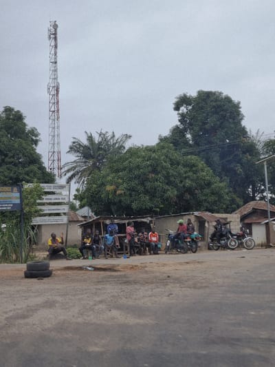 In Marke, Nigeria, a roadside scene unfolds under an overcast sky. A tall telecommunications tower dominates the left side of the frame, its metallic lattice structure reaching towards the sky. In front of a collection of simple, weathered buildings, a group of men are gathered. Some are seated on wooden benches or stools, while others stand near motorcycles. The men are casually dressed. Several motorcycles are parked alongside the buildings, some with riders already mounted. A signpost, likely indicating local government or development projects, stands to the left of the group. The ground in the foreground is a dusty, unpaved road. Stacked tires are visible on the left side of the road. Lush green trees and vegetation frame the scene, with the dense foliage of a large tree forming a backdrop behind the buildings and to the right. The overall impression is one of everyday life in a Nigerian town, with a focus on community gathering and transportation.