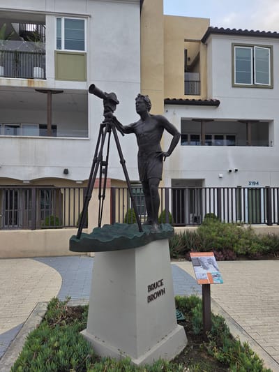 This is a medium shot, eye-level, outdoor photograph of a bronze statue of a man with a camera on a tripod, positioned in front of a modern apartment building. The statue is of Bruce Brown, a filmmaker, as indicated by the plaque at the base of the statue and a nearby information sign. The statue depicts Brown standing with his right hand on his hip, looking off to the left. He is wearing shorts and is bare-chested. The camera on the tripod is positioned to his left. The statue is mounted on a concrete pedestal with "BRUCE BROWN" inscribed on it. The building behind the statue is a multi-story structure with white and beige siding, balconies, and dark-trimmed windows. A black metal fence runs across the middle ground of the image. The foreground features a paved walkway and landscaping with succulents and other low-lying plants. The sky is overcast. The location is identified as the Bruce Brown Films Courtyard in Dana Point, California, United States of America.