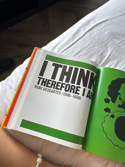 A book is open on a white surface, possibly a bed or sofa, with a person's arm visible in the lower right corner. The book is open to a page with large, bold black text that reads "I THINK THEREFORE I AM" followed by "RENE DESCARTES (1596-1650)". The left page has a green bar at the bottom. The right page is a bright green color and features a black graphic of a profile silhouette of a person's head with thought bubbles. The book has a red and yellow edge visible on the left side. The background is a blurred dark area. The scene is indoors.