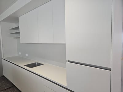 This media file shows a modern, minimalist kitchen in Darmstadt, Germany. The kitchen features sleek, white cabinetry with handleless doors and drawers, creating a clean and uncluttered aesthetic. The countertop is a light, neutral color, seamlessly integrating with the cabinets. A stainless steel sink is inset into the countertop, positioned to the left of the frame. Above the countertop, there are white wall cabinets, also with a minimalist design. To the far left, open shelving is visible, providing a contrast to the enclosed cabinets. The overall impression is one of sophisticated simplicity. The lighting suggests it is daytime, likely indoors with artificial light contributing to the bright, airy feel. There are no people visible in the scene. The location is identifiable as Darmstadt, Germany. There is no visible text in the image.