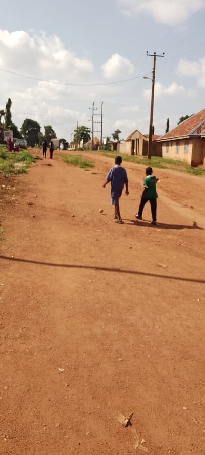 In Ajio, Nigeria, a wide dirt road stretches through a dusty, rural landscape under a bright, cloudy sky. Two young boys are walking away from the camera on the road. The boy on the left wears a blue t-shirt and striped shorts. The boy on the right wears a green t-shirt and black pants. The ground is a reddish-brown dirt, with scattered small stones and debris. To the left of the road, there is a patch of green grass and some vegetation. In the background, several small buildings with corrugated metal roofs are visible, along with utility poles and power lines. A few other people are walking further down the road, and a car is parked on the left side. The overall impression is a simple, unpaved street in a developing area.