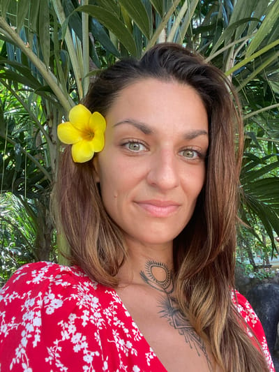A woman with light skin, brown hair with blonde highlights, and green eyes poses for a photo outdoors. A yellow flower is tucked behind her ear. She is wearing a red and white floral dress. Tattoos are visible on her neck and chest. Lush green foliage surrounds her.
The location is Dĩ An, Vietnam.