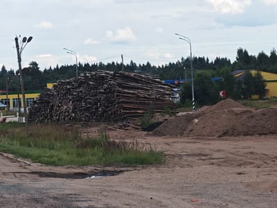 The image depicts an outdoor scene at what appears to be a logging or industrial area. A massive pile of logs dominates the center of the frame, stacked high and extending horizontally across the image. In the foreground, a dirt road with tire tracks and a small puddle is visible. To the left, a tall wooden pole with two streetlights is situated, with power lines extending from it. Beyond the logs, a dense forest forms the background, with a few buildings interspersed within it. One building on the left has a yellow facade with windows, and further to the right, another yellow building with a darker roof is visible. A blue sign with yellow lettering, possibly indicating a business name, is partially obscured by the logs. In the mid-ground, to the right of the logs, a large mound of earth or sand is piled up. The sky is overcast with scattered clouds. The overall impression is one of an active work or storage site in a semi-rural or industrial setting, likely on the outskirts of a populated area.