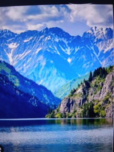 A body of water, likely a lake, is in the foreground. The water is calm with slight ripples and reflects the scenery. Beyond the water, a steep, rocky mountainside rises, covered in patches of green vegetation and scattered evergreen trees. In the distance, imposing snow-capped mountains dominate the horizon, their peaks sharp and dramatic against a sky filled with light, broken clouds. The mountains display varying shades of blue and white, indicative of distance and snow cover. The scene suggests a natural, mountainous landscape, potentially within or near the city of Auk, Kyrgyzstan, given its mountainous surroundings. There are no people or discernible actions visible in the image.