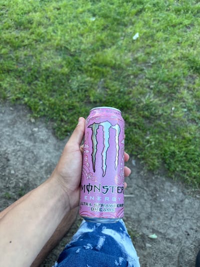 A right hand is holding a pink Monster Energy Ultra Strawberry Dreams can in an outdoor setting. The background consists of green grass and a gray concrete surface. The person is wearing blue jeans. The location, Hayredin, Bulgaria, cannot be verified from the image.