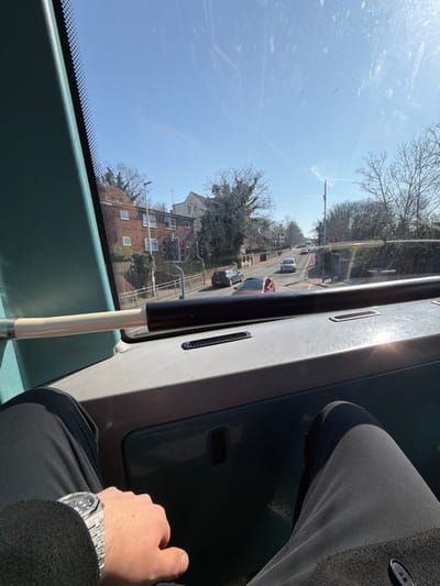 The image captures a sunny daytime street scene in Chatham, United Kingdom, viewed from the upper deck of a moving or stationary bus.
In the immediate foreground, the interior of the bus dashboard is visible, colored light grey, featuring two black rectangular ventilation grilles. A horizontal black handrail extends across the lower portion of the windshield. To the left, a teal-colored vertical structural pillar is prominent. The lower body of an individual, presumably the photographer, is partially visible; they wear black trousers and a black coat. Their right hand rests on their thigh, displaying a silver-colored wristwatch with a textured metallic band.
Beyond the bus windshield, a two-lane asphalt road, appearing to have a slight uphill gradient, dominates the midground. Several vehicles are present on the road, including a dark grey car followed by a red hatchback in the left lane, and a white van in the opposite lane further ahead. A traffic light on the left side of the road displays a red signal. Adjacent to the road on the left, a metal pedestrian railing is visible, behind which stands a retaining wall. Above the wall, a row of two-story brick residential buildings with white-framed windows is situated. The right side of the road is lined with bare deciduous trees and shrubs, indicative of winter or early spring.
The background shows the road curving into the distance, with more bare trees and glimpses of distant buildings under a clear, bright blue sky. Contrails from aircraft are faintly visible in the upper sky.