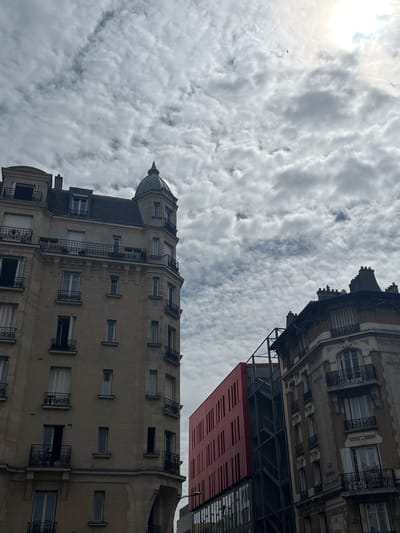 This media file shows an upward-angle view of two buildings against a cloudy sky. The building in the foreground is a light-colored, multi-story structure with a rounded corner and a small dome on top. It features numerous windows, many with black wrought-iron balconies. The building in the background is darker, with ornate architectural details at the top, including decorative cornices and small turrets. Below this upper portion, there is a section of red, horizontally-slatted material, possibly part of a modern addition or renovation.

The sky is filled with a dense layer of textured, greyish-white clouds. The sun is partially visible in the upper left corner, casting a bright, diffused light that suggests it is daytime, though the cloud cover obscures a clear view of the sky. No people are visible in the image. The scene appears to be an urban environment, given the presence of the buildings. The architecture suggests an older European city. There is no visible text. The overall impression is one of an overcast day in a city. The image is oriented sideways.