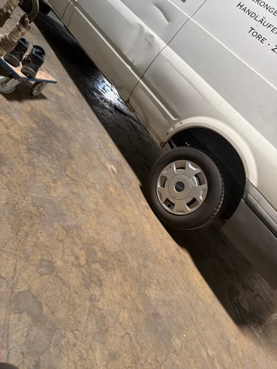 A pair of dark-colored sneakers is placed on a wooden dollies with four wheels. The dollies is positioned on a concrete floor in front of a white van. The van is parked diagonally in the scene, with its front left tire visible. The floor exhibits a cracked and stained pattern. In the background, text is visible on the side of the van, indicating it may be a commercial vehicle. The overall impression is one of a storage or workshop environment, possibly in an industrial area of Osnabrück, Germany.